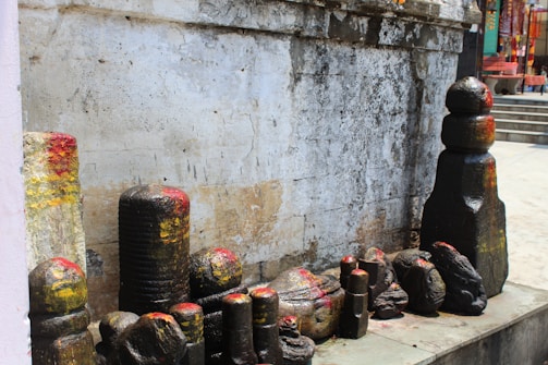 A collection of stone carvings and sculptures is placed against an old, weathered wall. The stones are various sizes and have colorful markings, primarily in red and yellow. Some of the formations appear to be shaped like traditional religious symbols or monuments.