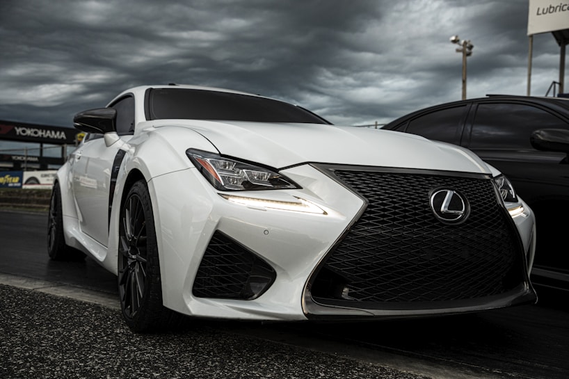 A sleek, white sports car is parked on a track, with a dynamic and angular design. The front grille is prominent, featuring a bold black mesh pattern, and the headlights are sharply styled. The sky above is overcast, adding dramatic contrast to the car's glossy surface. Additional details like the car's side mirrors and aerodynamic curves emphasize its high-performance capabilities.