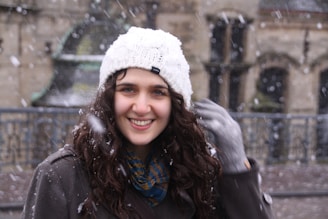 A smiling woman wearing a stylish winter coat, standing outdoors with soft snow falling.
