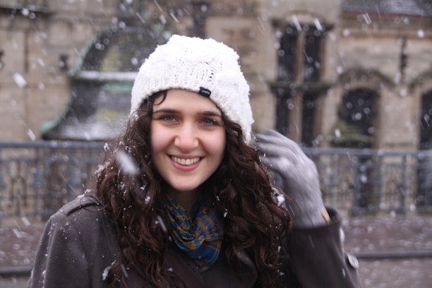 A smiling woman wearing a stylish winter coat, standing outdoors with soft snow falling.