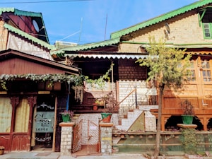 A rustic building with a combination of brick and wooden architecture is visible. The structure features a green, gabled roof with intricate woodwork. A small courtyard with stairs leads up to an entrance shaded by an awning. Potted plants and a tree add a touch of greenery to the scene. The signage on the left indicates it is named as a lodge. The building has been constructed with a mix of brown and beige hues, giving it an earthy appearance.
