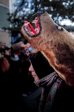 A person is wearing a bear costume that includes a detailed bear head with an open mouth, exposing sharp teeth and a bright red interior. The costume features intricate patterns, and the scene is set outdoors with blurred people in the background.