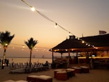A beach setting during sunset with palm trees and string lights illuminating a sandy area. There are wooden chairs and tables arranged on the sand, providing a relaxed atmosphere. In the background, a thatched-roof hut is visible, serving as a bar area with lights hanging from it. A silhouette of skyscrapers can be seen in the distance across the water.