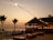 A beach setting during sunset with palm trees and string lights illuminating a sandy area. There are wooden chairs and tables arranged on the sand, providing a relaxed atmosphere. In the background, a thatched-roof hut is visible, serving as a bar area with lights hanging from it. A silhouette of skyscrapers can be seen in the distance across the water.