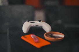 A gaming controller sits on a dark surface next to a vibrant orange smartphone and an ashtray containing a single burnt cigarette. The background is blurred, creating a cozy and relaxed atmosphere.