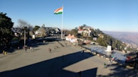A large open square with people walking around and an Indian flag flying on a tall pole. There are buildings and trees in the background, with hills visible in the distance.