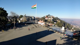 A large open square with people walking around and an Indian flag flying on a tall pole. There are buildings and trees in the background, with hills visible in the distance.