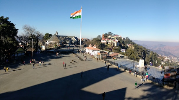A large open square with people walking around and an Indian flag flying on a tall pole. There are buildings and trees in the background, with hills visible in the distance.