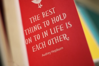 A red book cover features a quote in white text that reads 'The best thing to hold onto in life is each other.' The name Audrey Hepburn is displayed beneath the quote. The background is blurred with hints of yellow and teal colors.