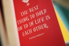 A red book cover features a quote in white text that reads 'The best thing to hold onto in life is each other.' The name Audrey Hepburn is displayed beneath the quote. The background is blurred with hints of yellow and teal colors.