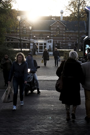 A group of people walking and riding a scooter towards a building with a sign that says 'MOOD'. The setting is outdoors in an urban area, with a road and brick paving. The lighting suggests late afternoon or early evening with a bright sun low in the sky, creating a strong backlight.