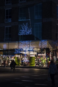 A storefront of a Rituals shop is shown during nighttime, illuminated by blue and white decorative lights shaped like trees. The glass windows of the store reflect the festive lights. People are walking in front of the store, and the interior is brightly lit, showcasing various products on shelves. The exterior of the building is dark, made of brick with several windows.