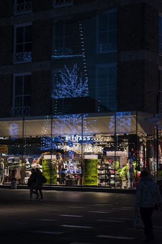 A storefront of a Rituals shop is shown during nighttime, illuminated by blue and white decorative lights shaped like trees. The glass windows of the store reflect the festive lights. People are walking in front of the store, and the interior is brightly lit, showcasing various products on shelves. The exterior of the building is dark, made of brick with several windows.