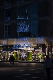 A storefront of a Rituals shop is shown during nighttime, illuminated by blue and white decorative lights shaped like trees. The glass windows of the store reflect the festive lights. People are walking in front of the store, and the interior is brightly lit, showcasing various products on shelves. The exterior of the building is dark, made of brick with several windows.