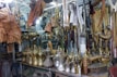 An assortment of traditional Arabic brass coffee pots, leather goods, and various wooden and metal artifacts are displayed in a cluttered vintage shop. The items are arranged on shelves and hanging from the ceiling, showcasing intricate designs and craftsmanship.