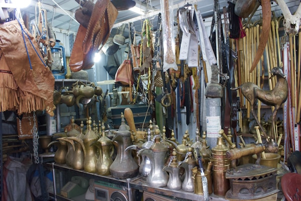 An assortment of traditional Arabic brass coffee pots, leather goods, and various wooden and metal artifacts are displayed in a cluttered vintage shop. The items are arranged on shelves and hanging from the ceiling, showcasing intricate designs and craftsmanship.
