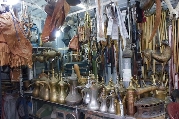 An assortment of traditional Arabic brass coffee pots, leather goods, and various wooden and metal artifacts are displayed in a cluttered vintage shop. The items are arranged on shelves and hanging from the ceiling, showcasing intricate designs and craftsmanship.