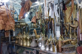 An assortment of traditional Arabic brass coffee pots, leather goods, and various wooden and metal artifacts are displayed in a cluttered vintage shop. The items are arranged on shelves and hanging from the ceiling, showcasing intricate designs and craftsmanship.