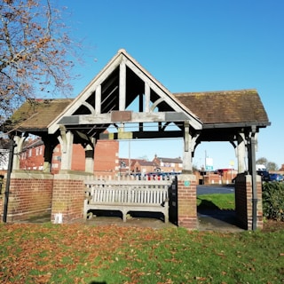 A covered wooden bench is situated within a small shelter made of wood and brick. The structure has an open triangular roof design. Surrounding the shelter are red brick buildings in the background, with a blue sky overhead. Autumn leaves and green grass cover the ground, while a partially bare tree is visible on the left side of the image.