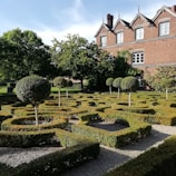 A meticulously landscaped garden featuring geometric hedges and topiary trees. The garden is in front of a traditional brick building with multiple windows and ornate roof details. The scene is bathed in sunlight, highlighting the greenery and architecture.