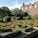 A meticulously landscaped garden featuring geometric hedges and topiary trees. The garden is in front of a traditional brick building with multiple windows and ornate roof details. The scene is bathed in sunlight, highlighting the greenery and architecture.