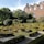 A meticulously landscaped garden featuring geometric hedges and topiary trees. The garden is in front of a traditional brick building with multiple windows and ornate roof details. The scene is bathed in sunlight, highlighting the greenery and architecture.