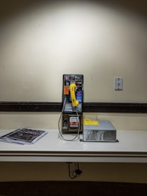 A wall-mounted payphone with a bright yellow handset is positioned on a shelf against a beige wall. To the right of the payphone is a metallic device with a yellow label on top, and to the left, there is a stack of newspapers. An electrical outlet is present on the wall above the shelf.
