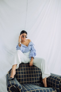 A person with sunglasses, wearing a blue and white striped off-shoulder top and white pants, sitting confidently on a plaid-patterned armchair. The background is a plain white fabric, creating a minimalist and fashionable setting.