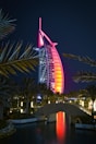 A sail-shaped luxury hotel is illuminated with vibrant red and pink lights against a dark night sky. Palm trees frame the scene, and a small stone bridge crosses over a calm body of water in the foreground, reflecting the colorful lights.