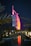 A sail-shaped luxury hotel is illuminated with vibrant red and pink lights against a dark night sky. Palm trees frame the scene, and a small stone bridge crosses over a calm body of water in the foreground, reflecting the colorful lights.