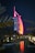 A sail-shaped luxury hotel is illuminated with vibrant red and pink lights against a dark night sky. Palm trees frame the scene, and a small stone bridge crosses over a calm body of water in the foreground, reflecting the colorful lights.