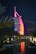 A sail-shaped luxury hotel is illuminated with vibrant red and pink lights against a dark night sky. Palm trees frame the scene, and a small stone bridge crosses over a calm body of water in the foreground, reflecting the colorful lights.