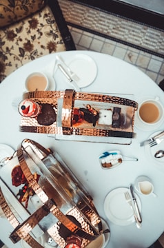 A round table set for tea, featuring a decorative arrangement with copper baskets and assorted pastries and treats. Cups of tea with saucers, cutlery, and small plates are organized neatly. The table is situated on a patterned tile floor, complemented by floral upholstery on nearby seating.