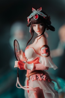 A detailed figurine of a character in traditional attire, featuring a decorative headpiece with a prominent red gemstone. The figure holds a circular fan adorned with red designs and wears a pink and white outfit embellished with floral motifs.