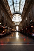 A grand shopping arcade with an impressive glass-domed ceiling, detailed architectural facades, and ornate decorations. The space is lined with upscale shops and cafes, where people are dining and walking.