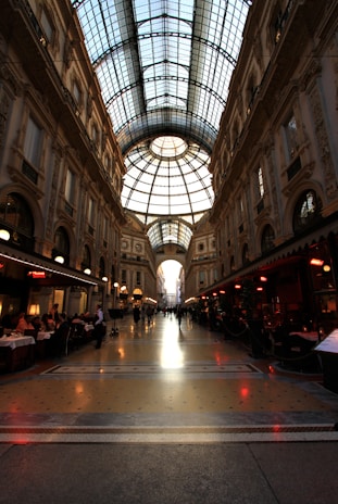 A grand shopping arcade with an impressive glass-domed ceiling, detailed architectural facades, and ornate decorations. The space is lined with upscale shops and cafes, where people are dining and walking.