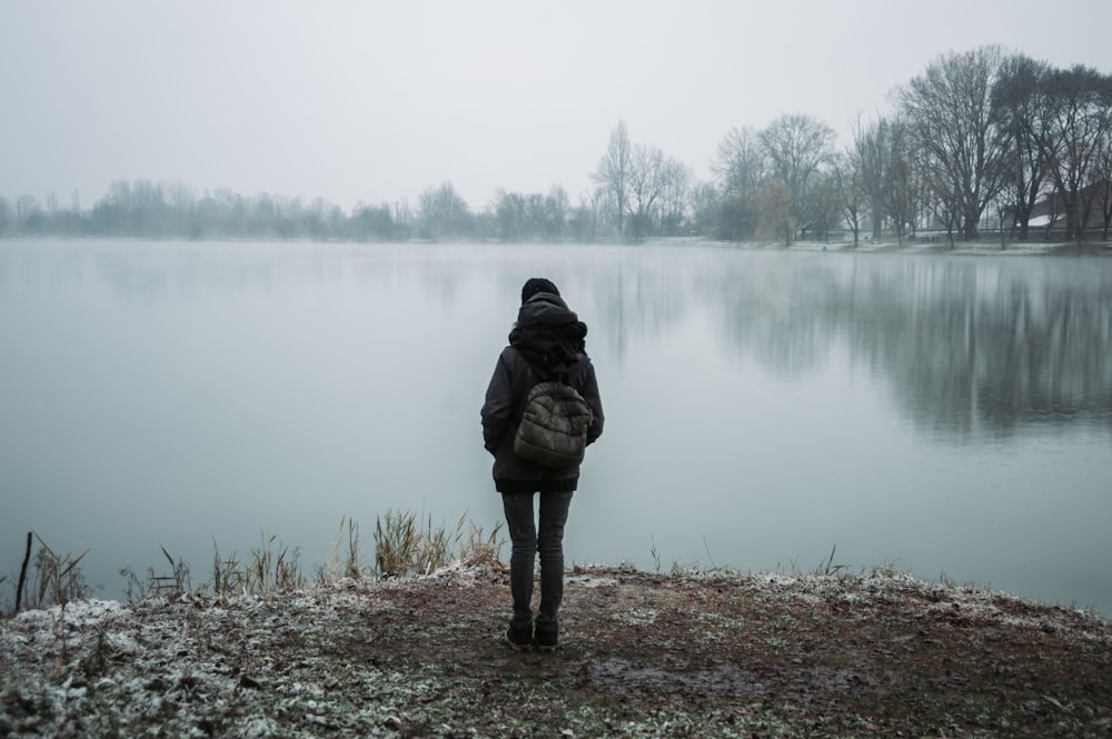 person wearing black hooded jacket standing and facing on body of water near trees in foggy day