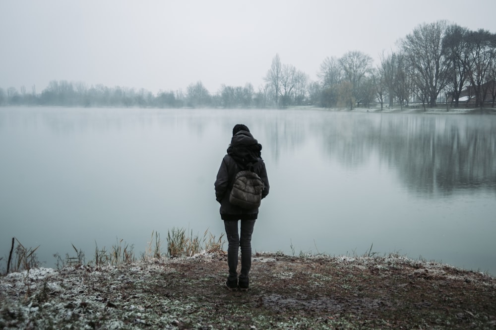 黒いフード付きのジャケットを着た人が、霧の日、木々の近くの水辺に立って向かい合っている