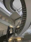 A spiral staircase with metal railings and concrete steps extends upwards in a modern building. The walls are made of glass blocks, allowing diffused light to fill the space. Fluorescent lights are mounted on the walls, adding illumination.