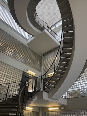 A spiral staircase with metal railings and concrete steps extends upwards in a modern building. The walls are made of glass blocks, allowing diffused light to fill the space. Fluorescent lights are mounted on the walls, adding illumination.