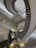 A spiral staircase with metal railings and concrete steps extends upwards in a modern building. The walls are made of glass blocks, allowing diffused light to fill the space. Fluorescent lights are mounted on the walls, adding illumination.