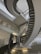 A spiral staircase with metal railings and concrete steps extends upwards in a modern building. The walls are made of glass blocks, allowing diffused light to fill the space. Fluorescent lights are mounted on the walls, adding illumination.