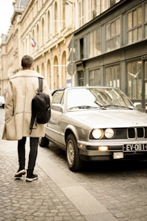 A person wearing a beige coat and carrying a black backpack walks on a cobblestone street next to a parked silver car. The street is lined with classic stone architecture, and a flag can be seen in the distance.