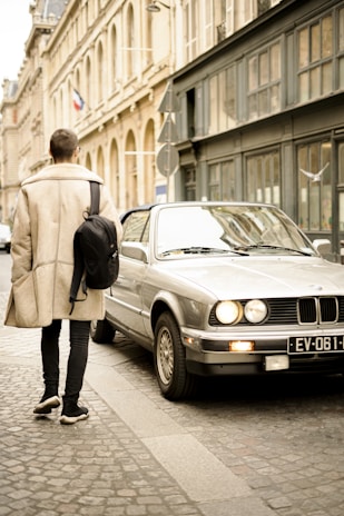 A person wearing a beige coat and carrying a black backpack walks on a cobblestone street next to a parked silver car. The street is lined with classic stone architecture, and a flag can be seen in the distance.