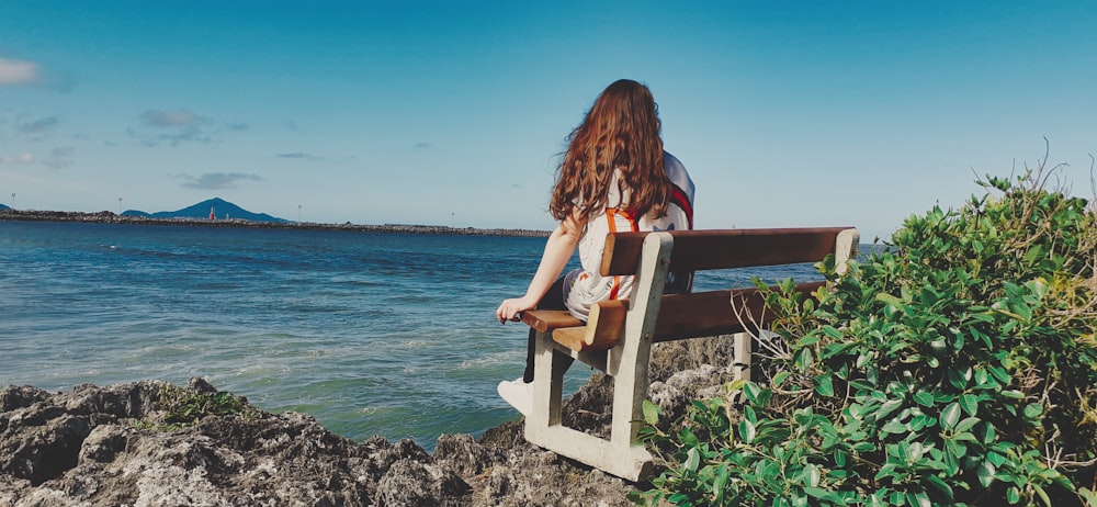 woman wearing white jacket sitting on white wooden bench facing body of water viewing mountain under blue and white sky