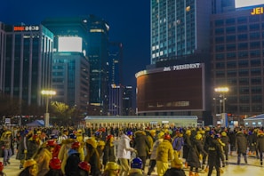 A bustling outdoor ice skating rink filled with people wearing colorful helmets and warm clothing. The background features tall, illuminated buildings with prominent signage, against a dark evening sky. Bright street lights are also visible, adding warmth to the scene.