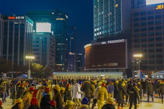 A bustling outdoor ice skating rink filled with people wearing colorful helmets and warm clothing. The background features tall, illuminated buildings with prominent signage, against a dark evening sky. Bright street lights are also visible, adding warmth to the scene.