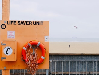 A beach scene with a prominently displayed life saver unit in orange, including a life preserver and a notice board with emergency instructions. In the background, a vast expanse of sandy beach leads to the sea where a distant person is flying a kite. The sky is slightly overcast, and a ship can be seen on the horizon.