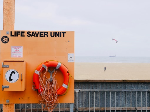 A beach scene with a prominently displayed life saver unit in orange, including a life preserver and a notice board with emergency instructions. In the background, a vast expanse of sandy beach leads to the sea where a distant person is flying a kite. The sky is slightly overcast, and a ship can be seen on the horizon.