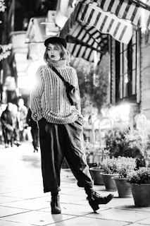 A person wearing a striped sweater and black beret walks confidently down a city street lined with potted plants and striped awnings. The atmosphere is urban and stylish, with a distinct black and white aesthetic.